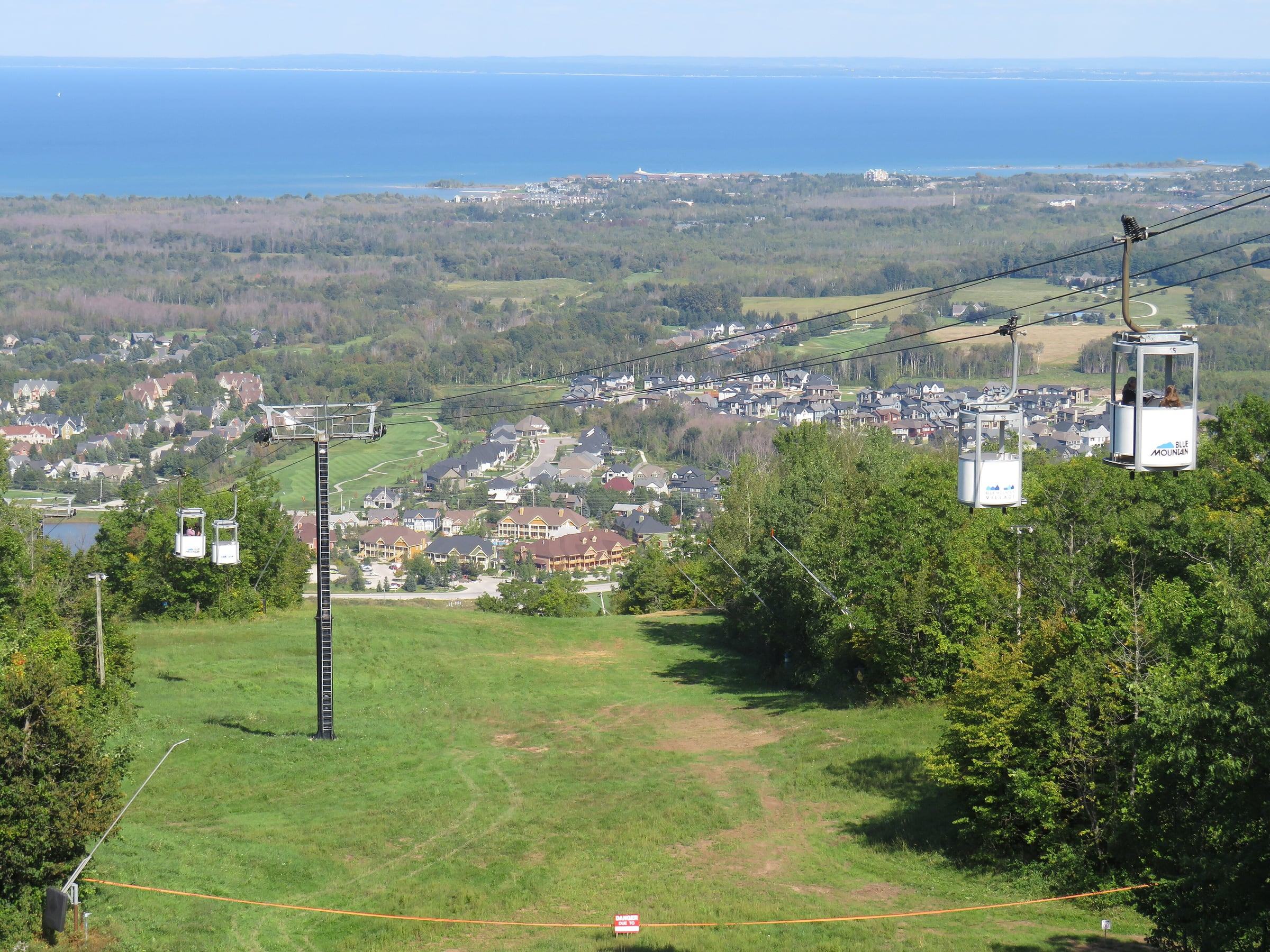 Blue Mountain gondola cabins above a grassy ski run, with Georgian Bay and Collingwood in the distance
