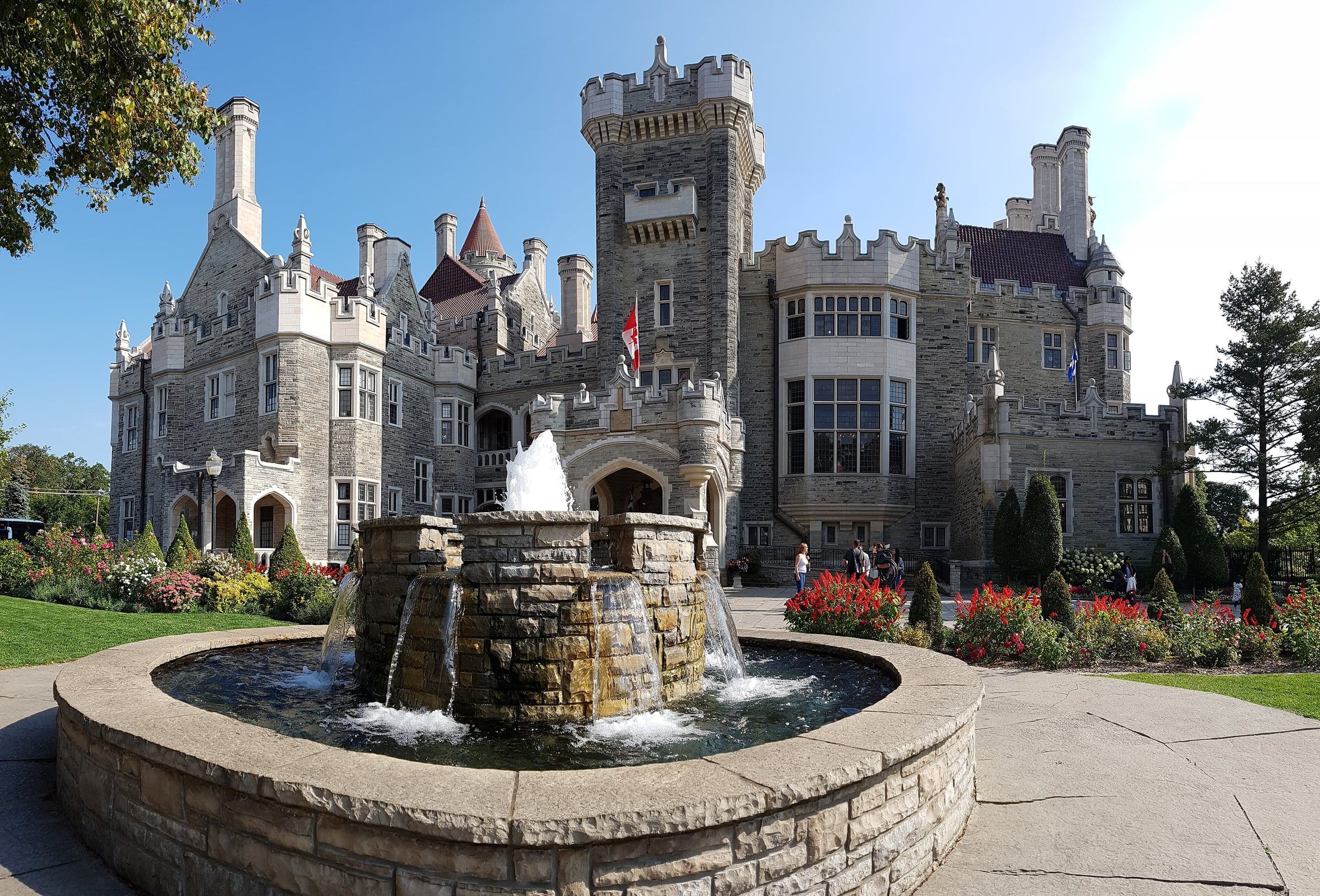 Casa Loma's stone towers and battlements behind a fountain and flower beds on a sunny day
